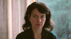 A person with dark hair sits in a domestic setting facing the camera. Their eyes look to something offscreen. The background is blurred, but shows, at left, a pastel coloured curtain. At right, we see a window through whichappears to be nature.