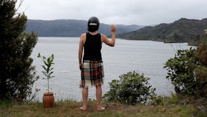 A person stands on the shoreline above a harbour. At their left is a potted plant, beside that is a naturally planted scrub bush. The person wear a tartan blanket, a black singlet and a black motorcycle crash helmet with the capital letters UDNRA printed on the back. Their right arm is raised as if in greeting or solidarity