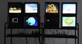 Eight vintage TV monitors are stacked in 2 x 2 configurations and displayed on black shelves with numerous wires and power cords visible. Each monitor shows a Hollywood movie studio logo.