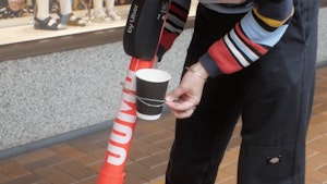 A close up of Bena’s hands whilst they tie an empty coffee cup to an electric scooter in Wellington CBD with a rubber band. The person is wearing a stripy jersey and black pants.
