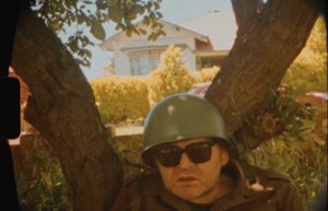 Wearing an army helmet and sunglasses, Ronnie sits against a tree with his mouth slightly open