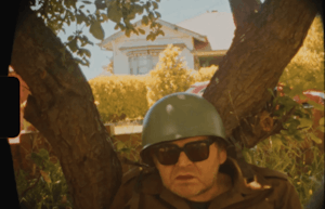 Wearing an army helmet and sunglasses, Ronnie sits against a tree with his mouth slightly open