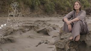 Emily sits barefoot on a barnacle-covered rock on the beach. Looking into the camera Emily looks peaceful and serious. There is an illustration of a character outlined in white sitting on the beach behind them.