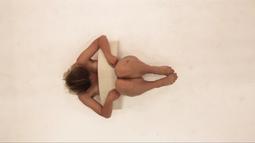 A birds-eye view of a person lying on a white floor with a semicircular sculpture across their torso.