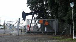 An oil well dredges behind large metal fences with warning signs attached. Parked cars are in the background.