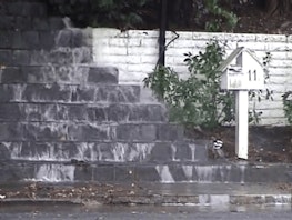 Water cascades down concrete steps as if there is a flood or leak, a mailbox with the number 11 on it stands up straight near the waterfall steps