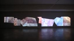 In a gallery abstract textured shapes of brown, grey and blue hues are projected on a wall.