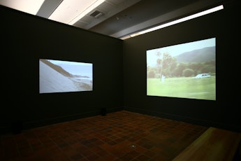 Two images projected into corners of a gallery, one showing a steep sand dune, the other a person in a white robe pointing a gun at a white passing car