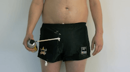 A topless man wearing short rugby shorts which are dripping with expanding foam, holds a can of expanding foam.