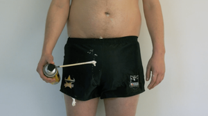 A topless man wearing short rugby shorts which are dripping with expanding foam, holds a can of expanding foam.
