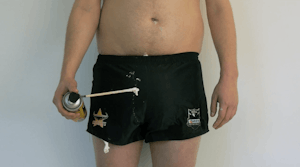 A topless man wearing short rugby shorts which are dripping with expanding foam, holds a can of expanding foam.