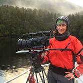 Alex Monteith is dressed in outdoors clothing standing near a large camera on a rig, they are standing next to a lake by a misty forest looking happy