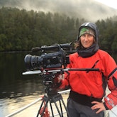 Alex Monteith is dressed in outdoors clothing standing near a large camera on a rig, they are standing next to a lake by a misty forest looking happy