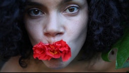 A person with curly black hair holds a red hibiscus flower in their mouth. Their gaze is directed at the camera.