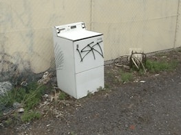 A discarded and graffitied washing machine sits near pieces of rubbish in a vacant car park or lot near a wire mesh fence.