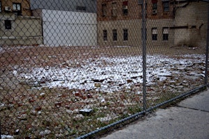 An area of rubble, behind a metal fence, covered in a white substance which could be snow. A brick building is in the background.