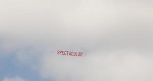 A large red ariel banner is flown through the air, towed from an out of the shot aeroplane. The text on the banner reads "SPECTACULAR." in large red lettering against a cloudy blue sky