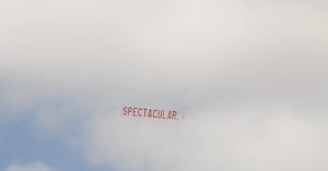 A large red ariel banner is flown through the air, towed from an out of the shot aeroplane. The text on the banner reads "SPECTACULAR." in large red lettering against a cloudy blue sky