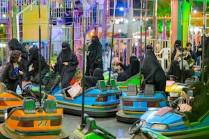 A group of people are getting into brightly coloured bumper cars.  Most of the people in the image are wearing burqa. Shot in a theme park in Abha, Saudi Arabia the work documents the life of Mahrajan Abha and the way that visitors to the fair negotiate a restrictive set of rules and regulations in the theme park which prohibits screaming on rides and ‘careless’ lifting of abayas.
