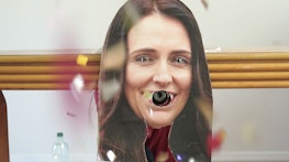 A cardboard cut-out of Jacinda Ardern has been attached to a wall. Jacinda's eyes and mouth have been cut out of the image, with a party popper coming through the hole where her mouth would be. Confetti is flying through the air as if the party popper has just gone off.