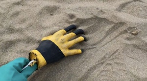 The artists hand with a rubber glove on, holds a sculpture of a hand also wearing a rubber glove. They hold the sculpture by a stick are raking sand with it.