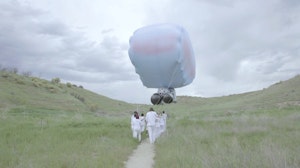 A group of people in white outfits carry the strings of a giant balloon through a grassy landscape