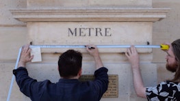 Two men hold up a tape measure to the base of a column on a public edifice in the Place Vendôme, Paris, that reads 'mètre'.