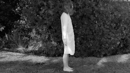 A black and white still shows Aliyah Winter standing outside on grass. Her arms are held out by her sides, extended slightly as if waiting for something or performing an action. The image is taken from a side profile perspective. She wears a long white dress shirt, standing on slightly wet grass in front of a hedge.