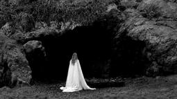 A person wearing a white sheet walks towards a large shadowy cave.