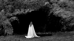 A person wearing a white sheet walks towards a large shadowy cave.