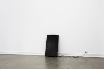 An LCD monitor with an almost imperceptably dark image stands against a wall, abjectly supported by a power cord plugged into the wall