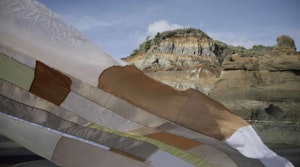 Flowing fabric flows gently with the wind in an outdoor location with a rock cliff behind it. The fabric piece is a patchwork made up of different beige coloured material, all with slightly different textures and transparency.
