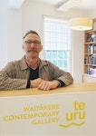 A person wearing glasses, standing behind a counter that says Te Uru Waitākere Contemporary Gallery