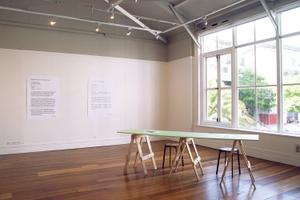 Bright light streams through large windows into a gallery with woooden floors, two large text images are on the wall and there is a table in the foreground
