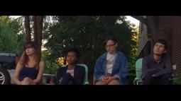 Four people sit in deck chairs looking at something. A house and tall trees stand behind them.