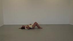 A person lies down in an empty white room with a bag of salt over their naked body.