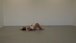 A person lies down in an empty white room with a bag of salt over their naked body.