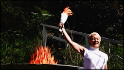 A person stands holding a victory torch made from paper beside a fire pit in which the fire is also made of paper.
