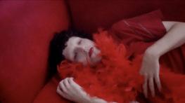 Zac wears red eye contacts, red lipstick, a red faux boa feather scarf, and a nose ring. They are lying down on a red couch, dark curly, short hair. The image is bright and hazy. Their hands delicately hold their feather boa whilst looking beyond the camera.