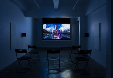 A screen play in a darkened room (blue hue): a man in traditional Samoan dress stands in front of a digital landscape of ocean and mountains.