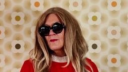 A person stands in front of a 60s wallpaper wearing a red dress, large black glasses, and a long blonde wig.