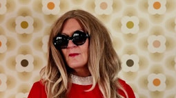 A person stands in front of a 60s wallpaper wearing a red dress, large black glasses, and a long blonde wig.