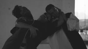 The black and white image shows three dancers leaning together with their bodies intertwined theatrically. The person in the middle wears sunglasses with large pointed studs.