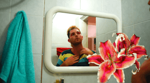 ZK gazes at themselves in a rounded mirror, they are wearing pink eye shadow and a striped top. Overlaid over the image are two large pink flowers