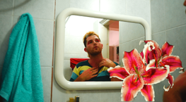 ZK gazes at themselves in a rounded mirror, they are wearing pink eye shadow and a striped top. Overlaid over the image are two large pink flowers