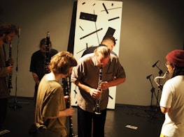 A group of people play woodwind instruments in a gallery. At centre rear, a large rectangular graphic score leans against the wall. On the floor are are two much smaller rectangles. All of the performers look down as they play, as if referencing the rectangles on the floor, which act as graphic scores.