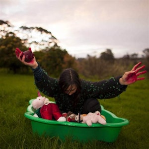 A person raises blood red hands to the sky whilst sitting in a green bucket in a field