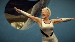 A smiling person wearing a silver leotard and silver arm bands dances with their arms outstretched. There is a large blue sculpture behind them.