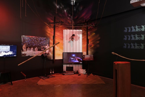 A dense multi-media installation including TV monitors, a pair of hanging sneakers, a computer, Pasifika musical instruments, stencil on the wall and more.