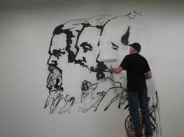 A person stands on a chair to paint over portraits of three figures painted in black paint.